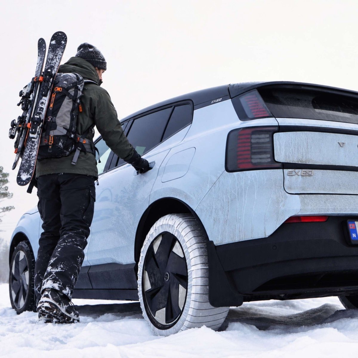 A snowy outdoor scene showing a person in winter gear standing beside a light blue electric vehicle that has NORSOXX snow socks mounted