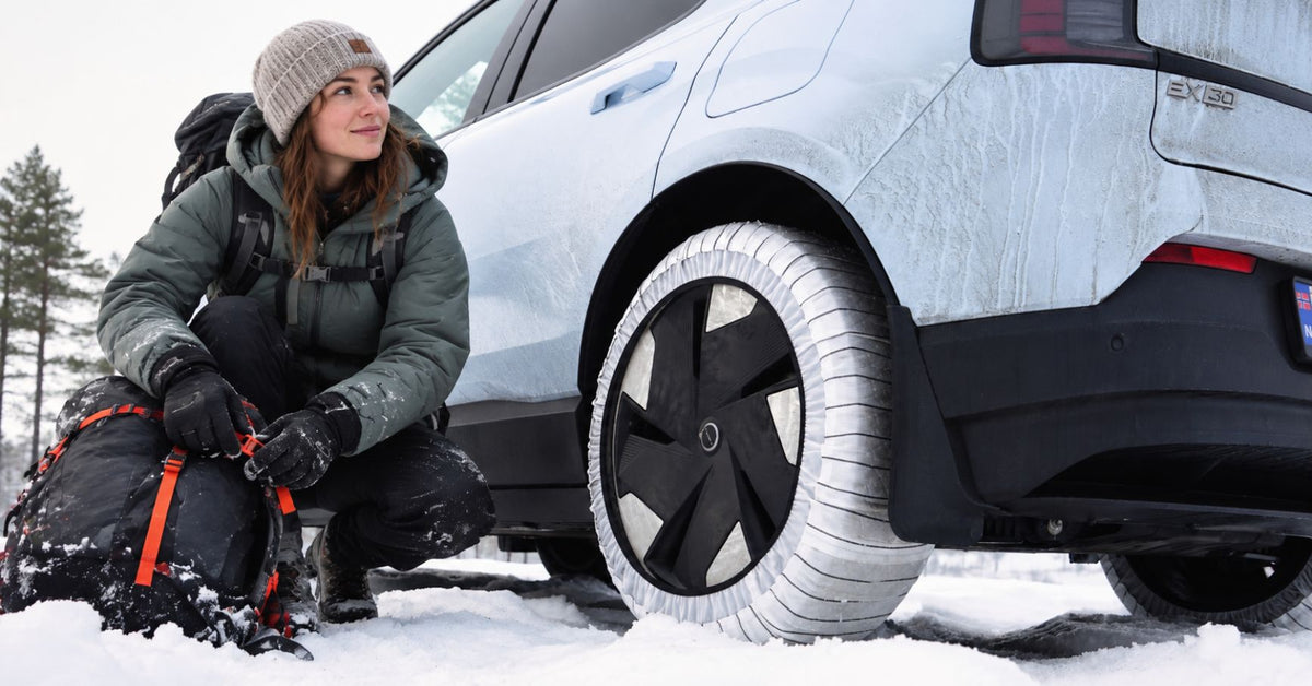 A woman in winter clothing kneels beside a white electric vehicle in the snow, showcasing snow socks mounted on the wheels of the car