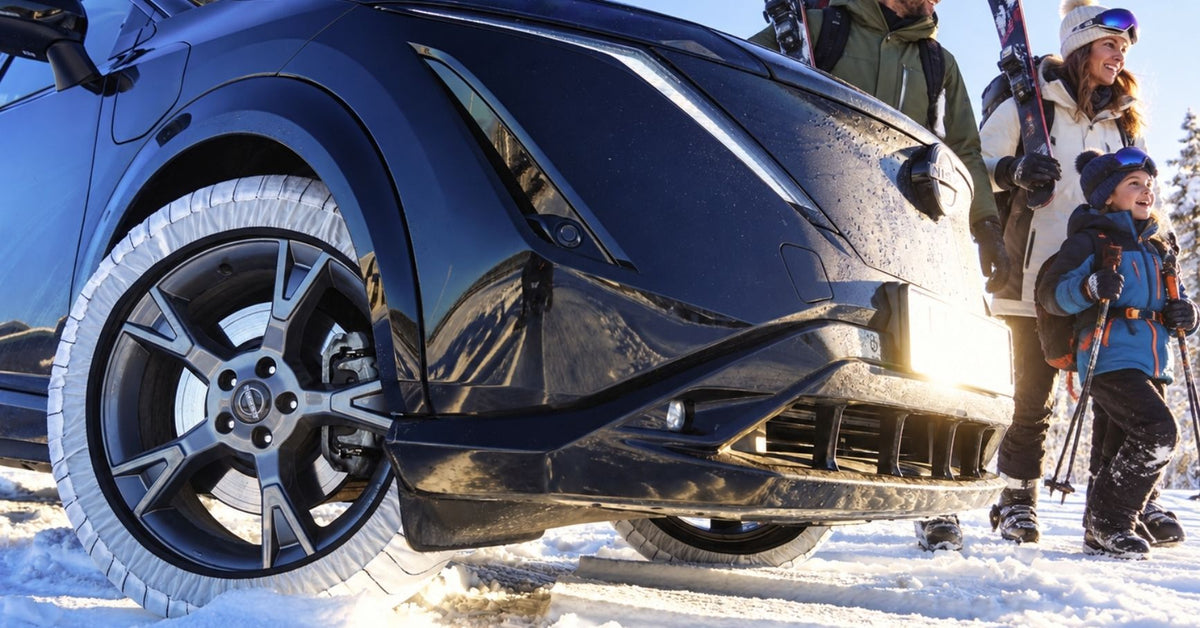 A close-up outdoor winter shot of the front wheel and bumper of a dark blue SUV in snow and snow socks mounted, with a family in winter clothing standing beside it holding skis.